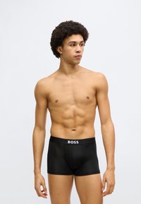 Fekete boxernadrág sima anyagból, logós derékpánttal. A modell egyenesen áll, kiemelve a testhezálló szabást és az atletikus alakot.