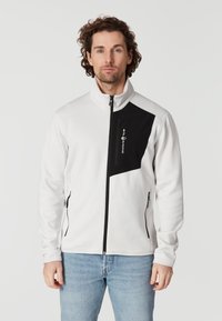 Vit zip-up jacka med svart accentpanel, med hög krage, sidofickor och en logo på bröstet. Slät, något texturerad tyg.