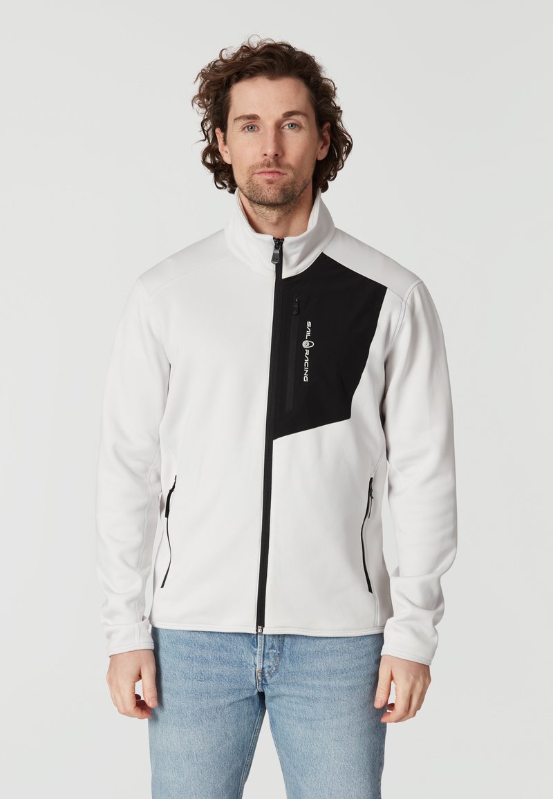 Vit zip-up jacka med svart accentpanel, med hög krage, sidofickor och en logo på bröstet. Slät, något texturerad tyg.