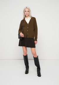 Cardigã verde-oliva com botões, camisa branca com gola, saia castanho escuro e botas pretas até ao joelho, combinados com uma pequena mala preta.