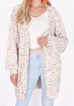 Femme portant un long cardigan en tricot blanc épais avec des taches multicolores, un jean bleu clair déchiré, un haut blanc, une ceinture beige et des boucles d’oreilles orange.