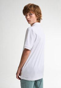 T-shirt polo blanc à manches courtes, avec un col et une coupe droite, associé à un short vert clair. Présente un petit logo sur l'ourlet.