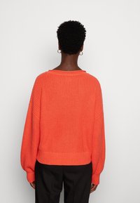 Krátký svetr s krátkými rukávy v jasně korálovém úpletu. Má široký výstřih a žebrované manžety a spodní lem. Zobrazen zezadu, nošený s černými kalhotami.