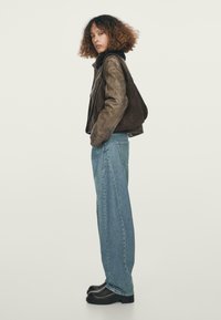 Brązowa skórzana kurtka, niebieskie szerokie dżinsy z denimu, czarne masywne buty. Model stoi bokiem na jasnym tle.