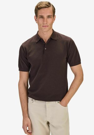 Nuori mies, jolla on yllään tummanruskea teksturoitu poolopaita ja vaaleanbeiget housut, seisoo oikea käsi taskussa yksinkertaisen taustan edessä.