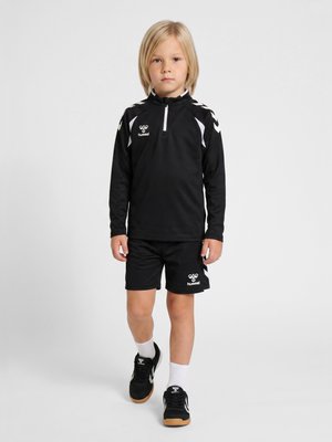 Barn med blondt hår iført sort sportstøj, sorte shorts, hvide sokker og sorte sneakers, der står på en ensfarvet lys baggrund.