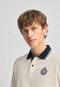 Beige Poloshirt mit einem navyfarbenen Kragen, ausgestattet mit einem Logo auf der linken Seite, gefertigt aus einem strukturierten Stoff und mit kontrastierenden Nähdetails.