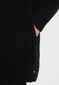 Schwarzer, gestrickter Pullover mit strukturiertem Muster, langen Ärmeln, Seitentaschen und einem gerippten Saum mit einem kleinen Logodetail.
