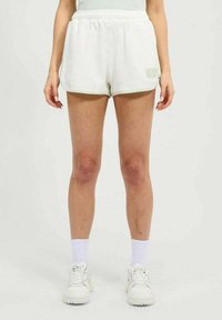 Mujer de pie vestida con pantalones cortos de tela acanalada blanca, calcetines blancos y zapatillas gruesas blancas sobre un fondo claro y liso.