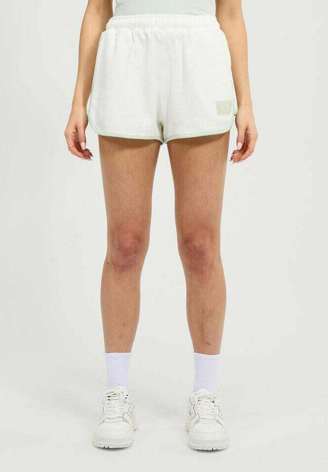 Mujer de pie vestida con pantalones cortos de tela acanalada blanca, calcetines blancos y zapatillas gruesas blancas sobre un fondo claro y liso.