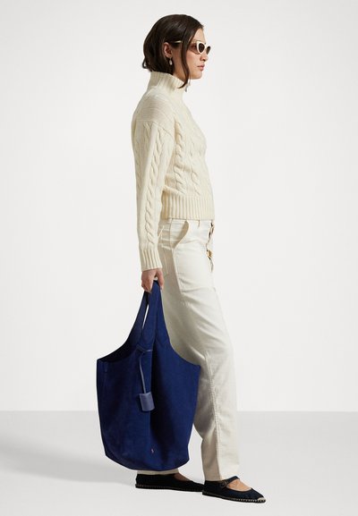 Tote bag en daim marine avec de longues poignées. Le modèle porte un pull texturé crème et un pantalon beige clair, complétant le look avec des ballerines noires.