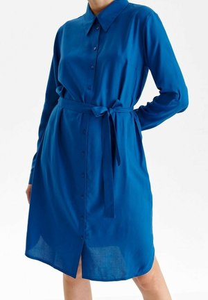 Kobieta ubrana w niebieską, sięgającą do kolan sukienkę na guziki z długim rękawem i paskiem w talii, stojąca na tle jednolitego, białego tła.