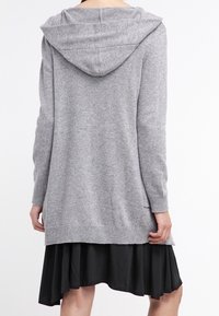 Cardigan largo gris con capucha, puños acanalados y aberturas laterales, hecho de un material de punto suave, combinado con un vestido negro acampanado debajo.