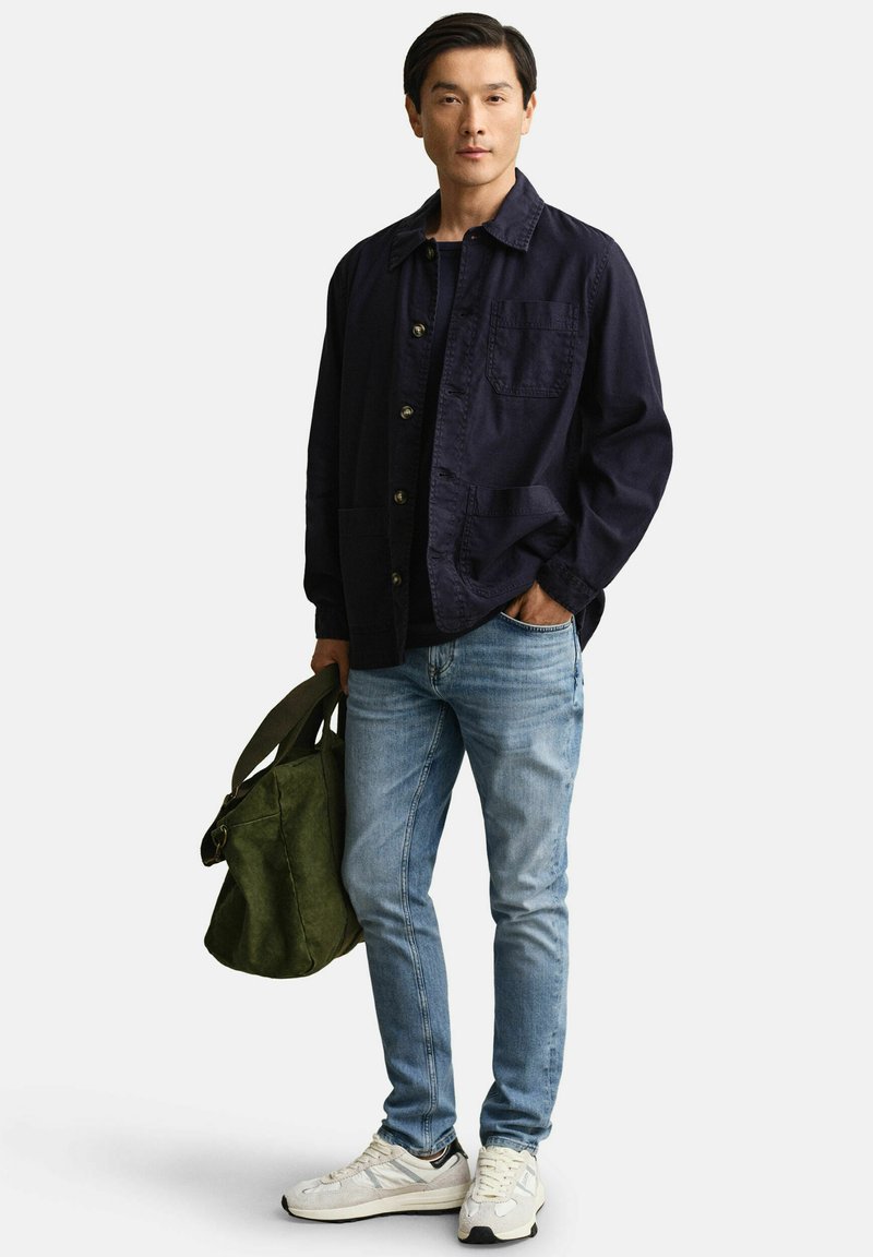 Hombre con chaqueta azul marino abotonada y vaqueros azul claro, de pie sosteniendo una bolsa verde oliva en una mano, con la otra mano en el bolsillo.