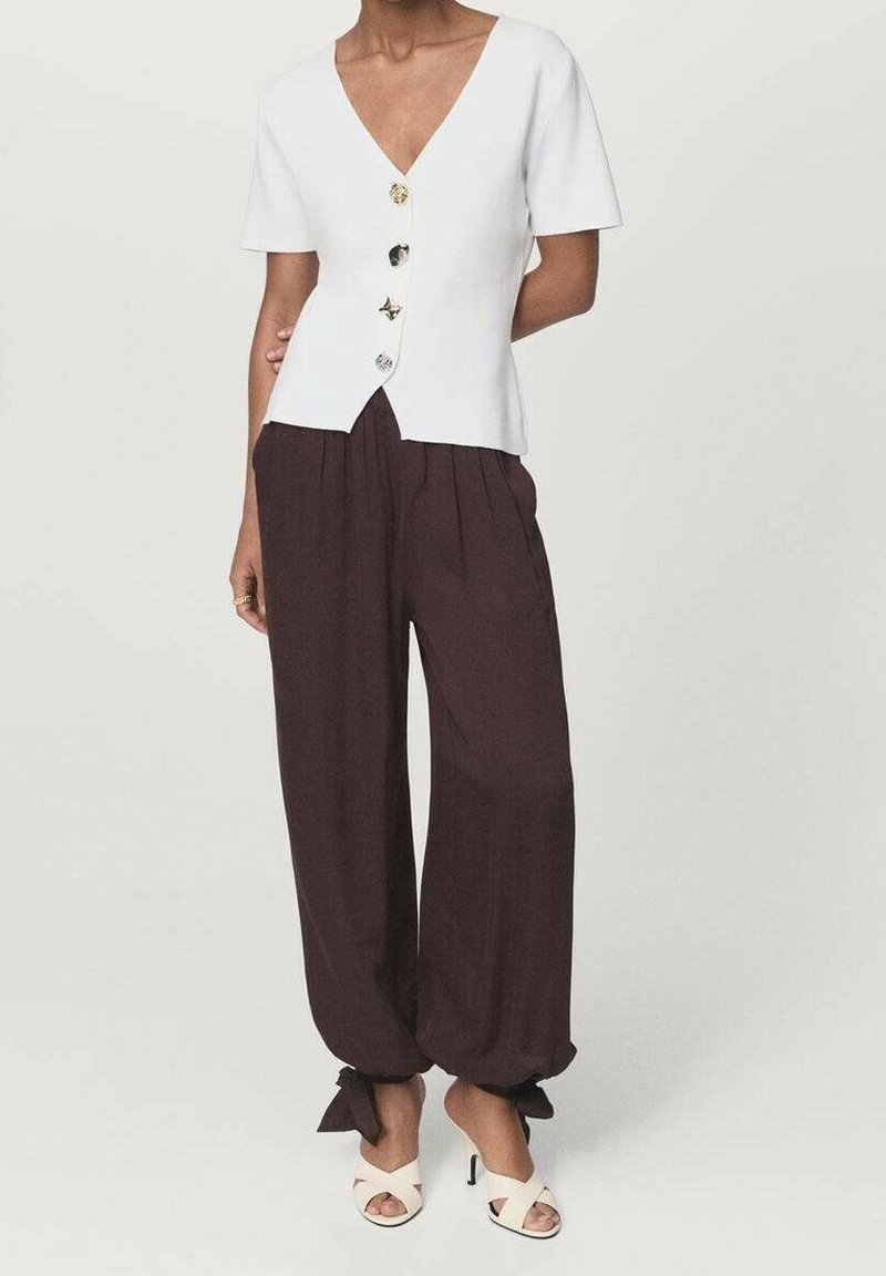 Femme portant un haut blanc à manches courtes boutonné, un pantalon ample marron foncé noué aux chevilles et des sandales blanches à talons ouverts, debout devant un fond uni.