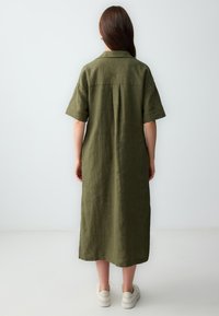 Olijfgroene linnen jurk met korte mouwen, een kraaghalslijn en een relaxte pasvorm, die tot halverwege de kuit reikt met een middelste rugplooi.