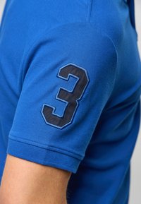 Modrá polo košile s texturovanou tkaninou, s ohrnutými rukávy a černým vyšitým číslem "3" na levém rukávu.