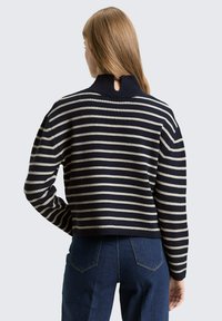 Pull rayé marine et crème avec un design court, col côtelé et détail œillet dans le dos. Texture tricotée et coupe décontractée. Pantalon en denim visible.
