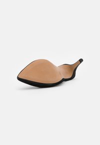 Chaussure noire à talon haut et bout pointu, posée sur le côté, montrant une semelle beige et une fine pointe de talon sur fond blanc.