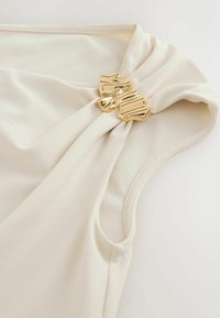 Cream fabric sleeveless top with a twisted neckline secured by a shiny gold decorative brooch.