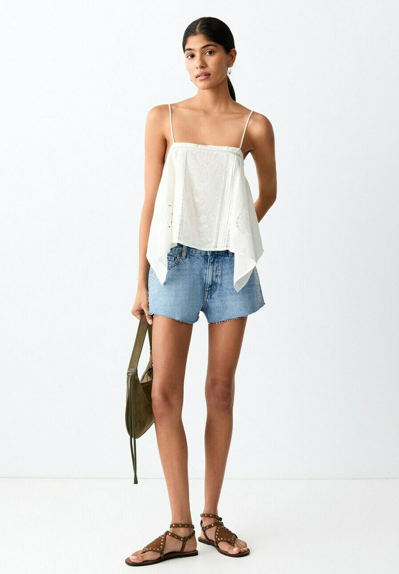 Jeune femme portant un haut blanc sans manches, un short en jean, des sandales marron cloutées, tenant un sac vert olive sur fond blanc.