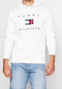 Sudadera blanca hecha de una mezcla de algodón, con un bolsillo frontal, capucha ajustable y un logo impreso con un diseño de bandera rojo, blanco y azul.