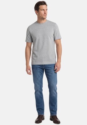 Mężczyzna stojący, ubrany w prozny jasnoszary T-shirt z krótkim rękawem, niebieskie dżinsy i ciemnobrązowe buty, patrzący w prawo.