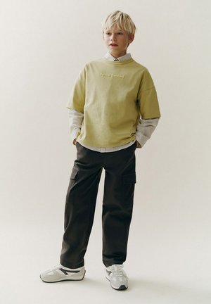 Garçon blond debout les mains dans les poches, portant une chemise beige superposée, un t-shirt vert olive, un pantalon cargo noir et des baskets blanches, sur un fond uni.