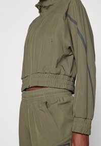 Olive green athletic jacket with a cropped design, elastic hem and cuffs, zippered pockets, and gray side stripes. Smooth fabric texture.