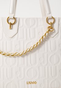 Borsa a tracolla bianca con texture, logo in rilievo, dettagli in catena dorata e forma rettangolare. Presenta doppi manici e accessori in metallo.