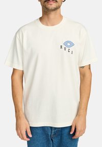 Krémové bavlněné tričko s krátkým rukávem, s modrou grafikou oka a černým textem "RVCA" na levé části hrudníku. Volný střih.