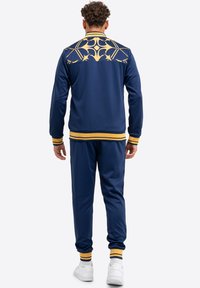 Survêtement bleu marine avec un design graphique doré au dos, agrémenté d'accents rayés jaunes et noirs aux poignets et à la taille.