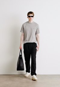 Grå bomulls T-skjorte med korte ermer, svarte lette bukser, hvite joggesko og en svart totebag. Modellen har på seg mørke solbriller.
