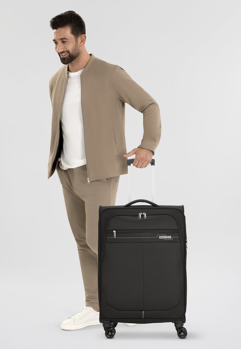 Valigia nera con ruote, dotata di chiusure a zip, una tasca frontale e quattro ruote girevoli; realizzata in un tessuto resistente, presenta un design elegante.