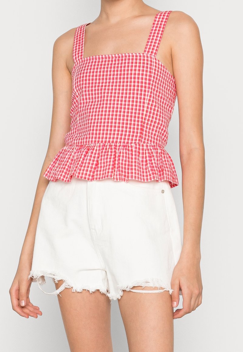 Haut peplum à carreaux vichy rouges et blancs avec bretelles carrées et ourlet évasé, associé à un short blanc déchiré avec des bords effilochés.