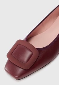 Burgundy leather flat shoe with a square rubber buckle detail, rounded toe, and smooth inner lining. Rubber sole for grip.