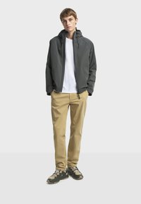 Graue Jacke mit Kapuze, Reißverschluss vorne und Seitentaschen, kombiniert mit einem weißen Hemd und khakifarbenen Hosen. Die Schuhwerk hat dunkle Akzente.