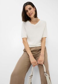 Lys beige kortærmet sweater med V-hals, parret med brune vide bukser. Modellen holder en lysvævet håndtaske.