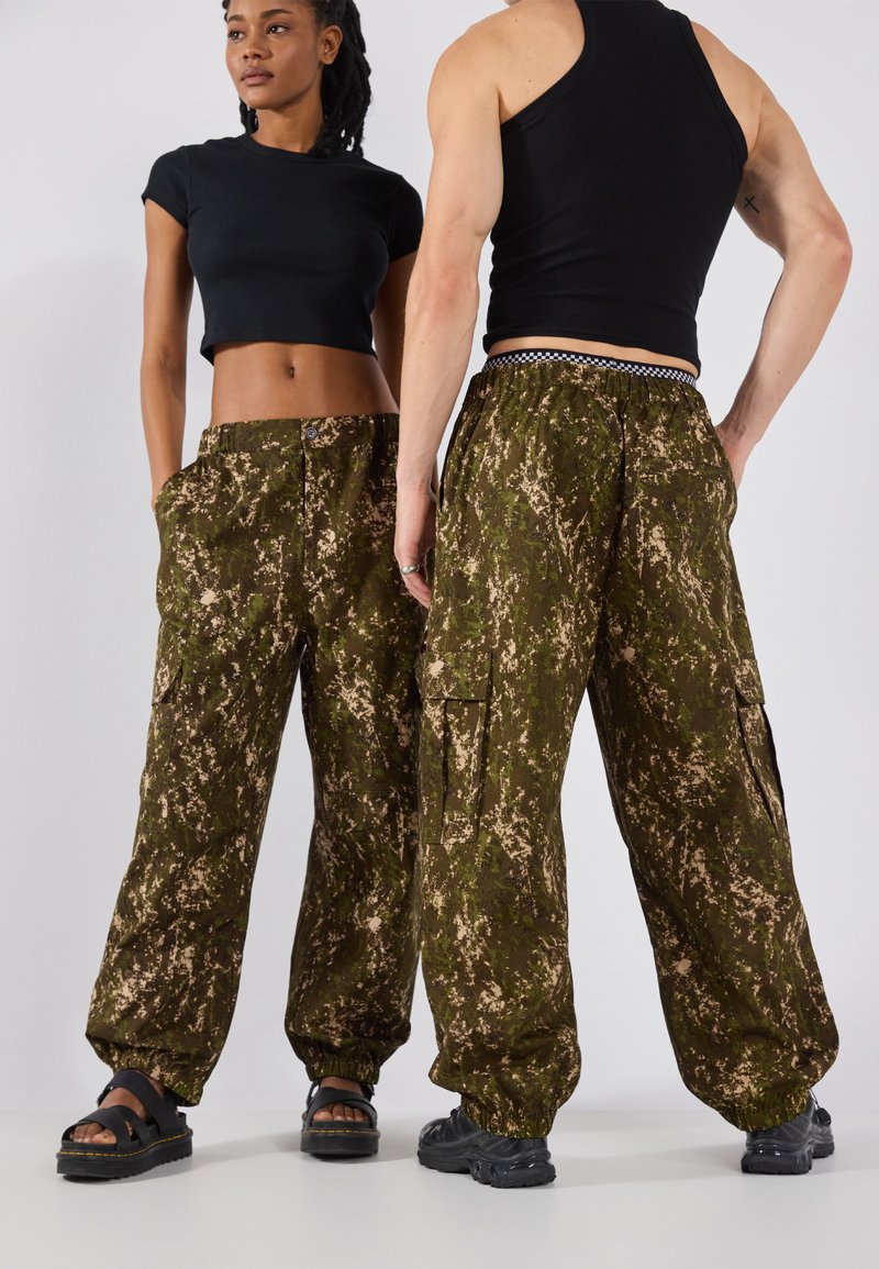 Cargo-Hosen in einem grün-beigen Camouflage-Muster, mit Seitentaschen, elastischen Bündchen und lockerer Passform. Getragen mit enganliegenden schwarzen Oberteilen.