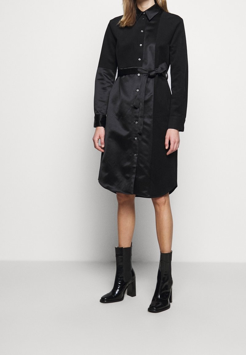 Svart skjortklänning med satinfinish, lång ärm, knappstängning fram och bälte i midjan. Har en något rundad nederkant och manschetter.
