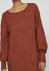 Pull en tricot couleur rouille avec une coupe décontractée, un large col et des manches volumineuses. Le tissu est doux au toucher et les poignets sont côtelés.