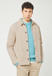Beige canvas jacket with black buttons, two front pockets, worn over a light blue textured sweater. Neutral trousers complete the outfit.