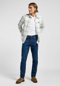 Ljus rutig skjorta med blå och beige nyanser, bärs över en vit t-shirt. Mörkblå jeans och bruna mockaloafers fulländar looken.