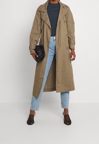 Kahakitränckåp med dubbelknäppning, bärs över en blå ribbad topp och ljus tvättade jeans. Svart clutch och öppna nätklackar ingår.