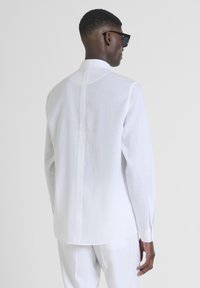 Camicia bianca in lino con maniche lunghe, colletto alto e cucitura centrale sul retro. Texture liscia e vestibilità sartoriale, abbinata a pantaloni di colore chiaro.