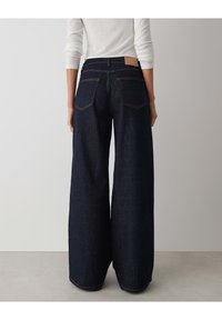 Wide-leg dark denim jeans with a high waist, visible stitching, and two back pockets. Features a tan leather patch on the waistband.