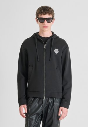 Sudadera negra con cremallera y capucha ajustable, logo bordado en el pecho y bolsillos delanteros. Usada con pantalones negros brillantes y gafas de sol.