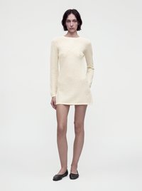 Femme aux cheveux courts et foncés portant une robe courte en maille crème à manches longues et des chaussures plates noires, debout devant un fond blanc uni.