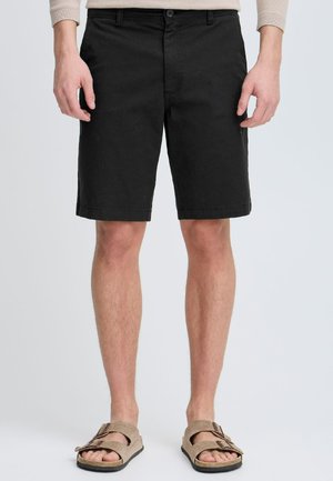 Bermuda nera in cotone con taglio dritto, dotata di tasche frontali e chiusura a bottone. Abbinata a sandali color cammello.