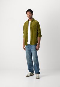 Cămașă din fâș de culoare verde măsliniu peste un tricou alb, asortată cu blugi și adidași verde deschis cu accente maro. Modelul stă cu încredere.